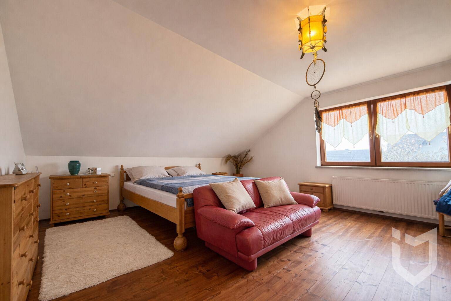 Schlafzimmer im Dachgeschoss mit Holzboden, Doppelbett und rotem Sofa unter schräger Decke.