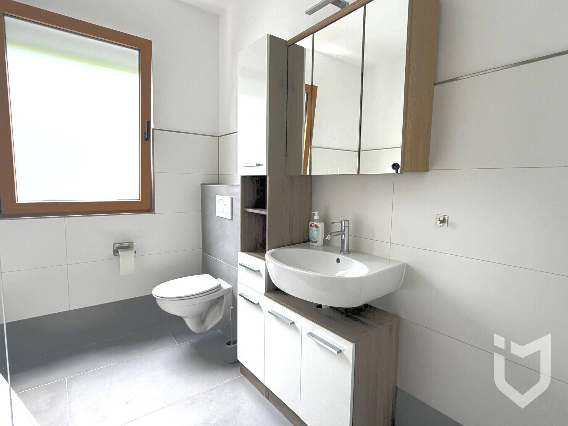 Helles Badezimmer mit wandhängendem WC, Einzelwaschbecken, Spiegelschrank und einem Fenster.