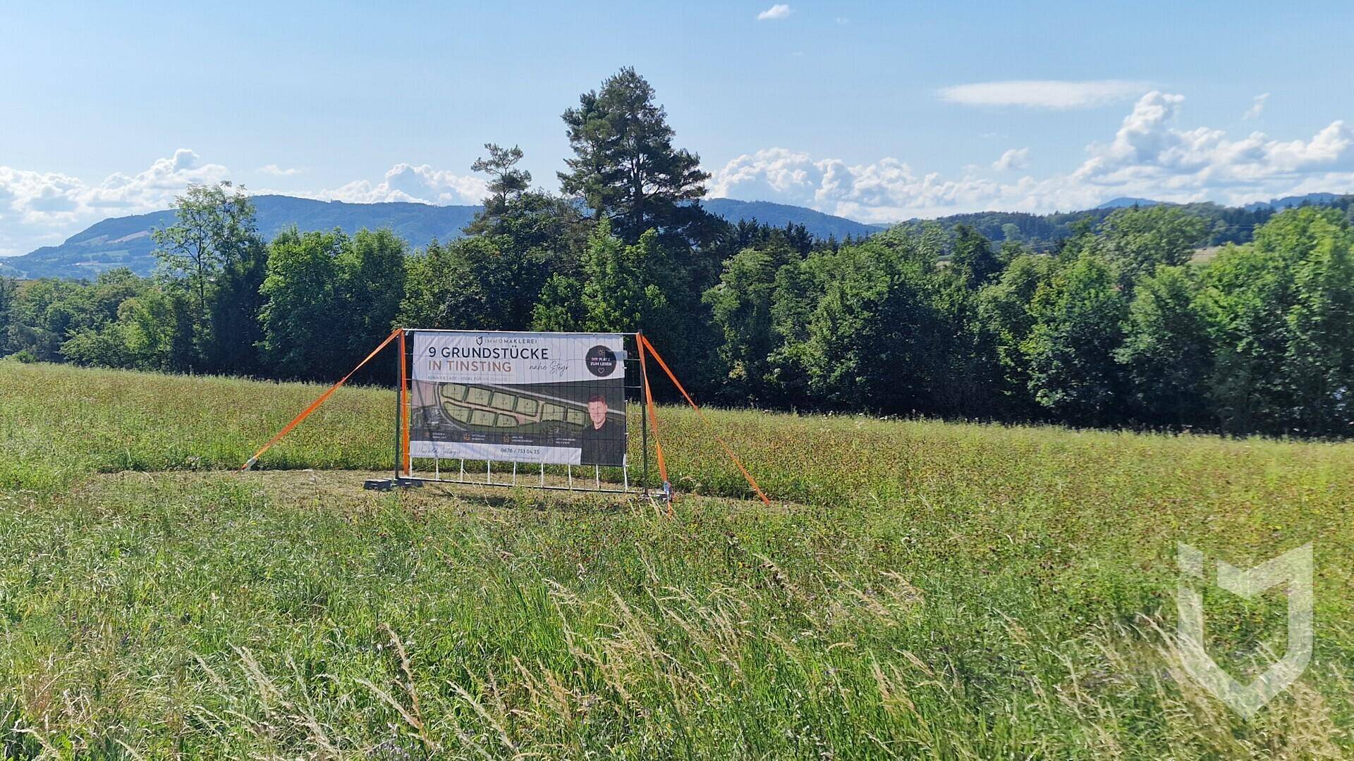 Weitläufiges Feld mit einem Hinweisschild auf Grundstücke, umgeben von Bäumen und Hügeln unter blauem Himmel.