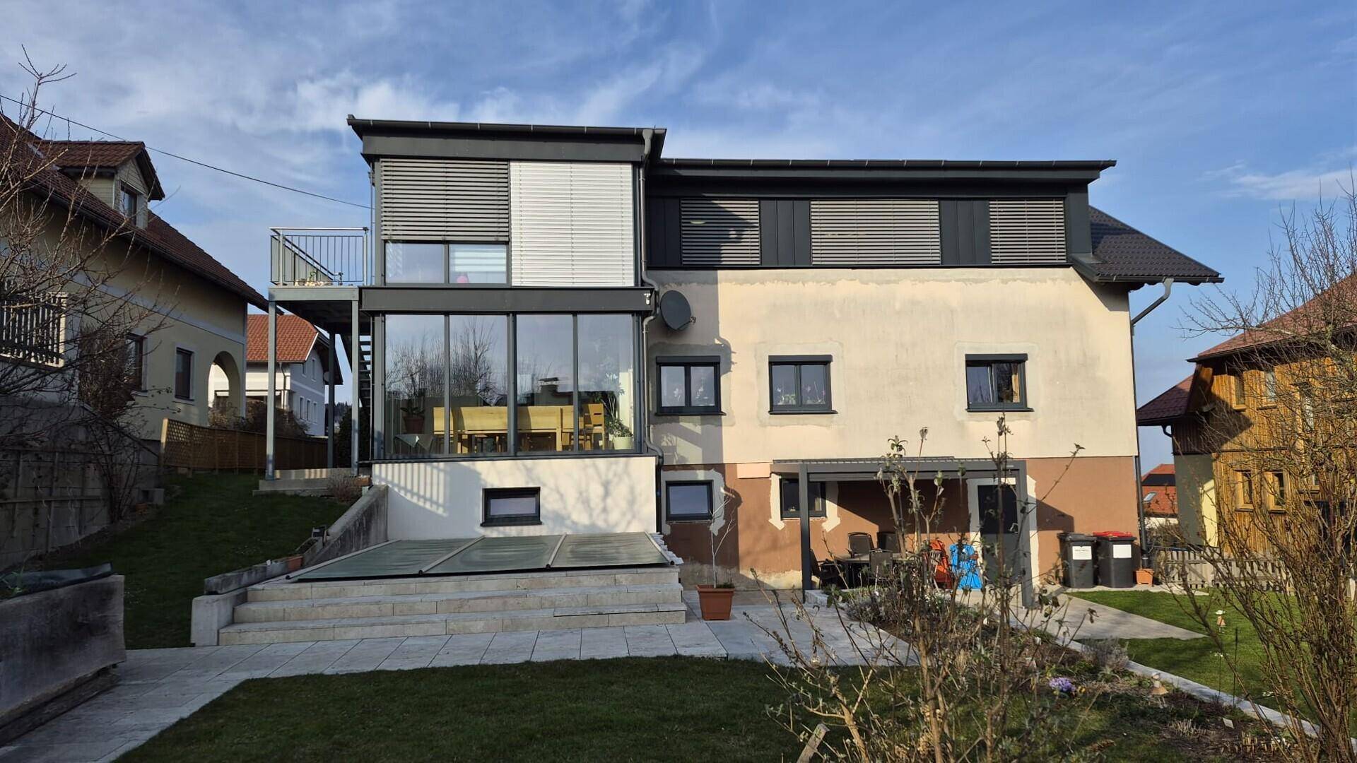 Rückansicht des Hauses mit Garten und Poolbereich, umgeben von ländlicher Umgebung bei bewölktem Himmel.