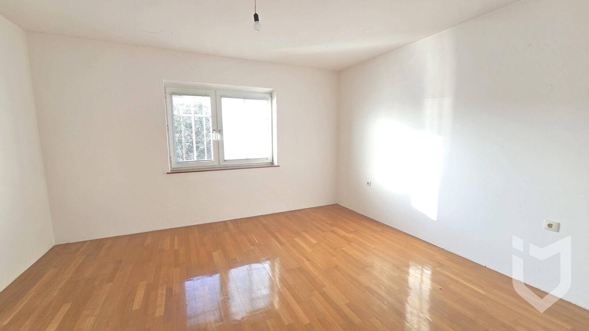 Leeres Zimmer mit glänzendem Parkettboden und einem Fenster, das viel natürliches Licht hereinlässt.