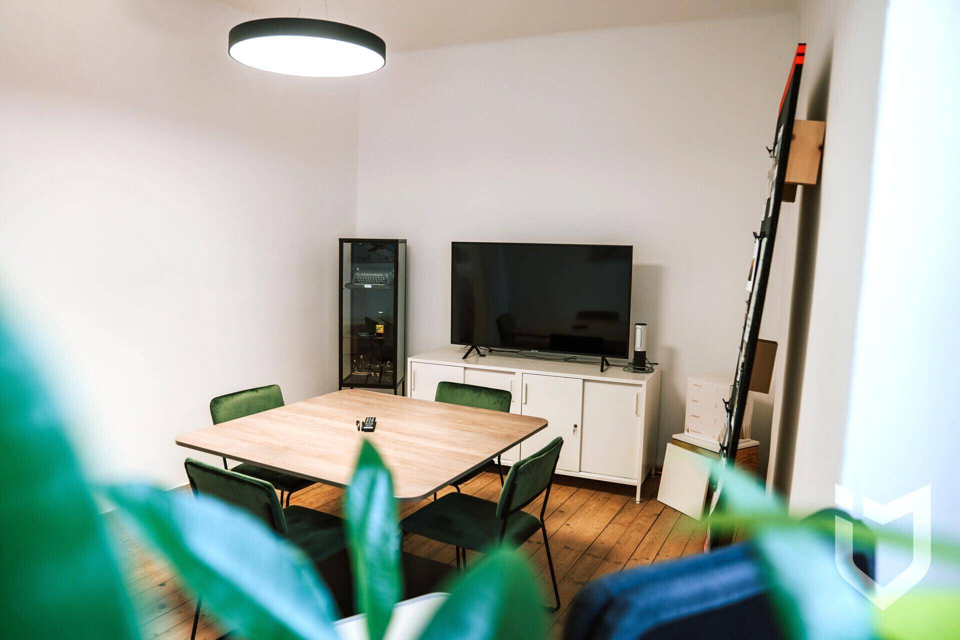 Gemütlicher Lounge-Bereich mit Holztisch, grünen Stühlen, großem Fernseher und einem modernen Sideboard.
