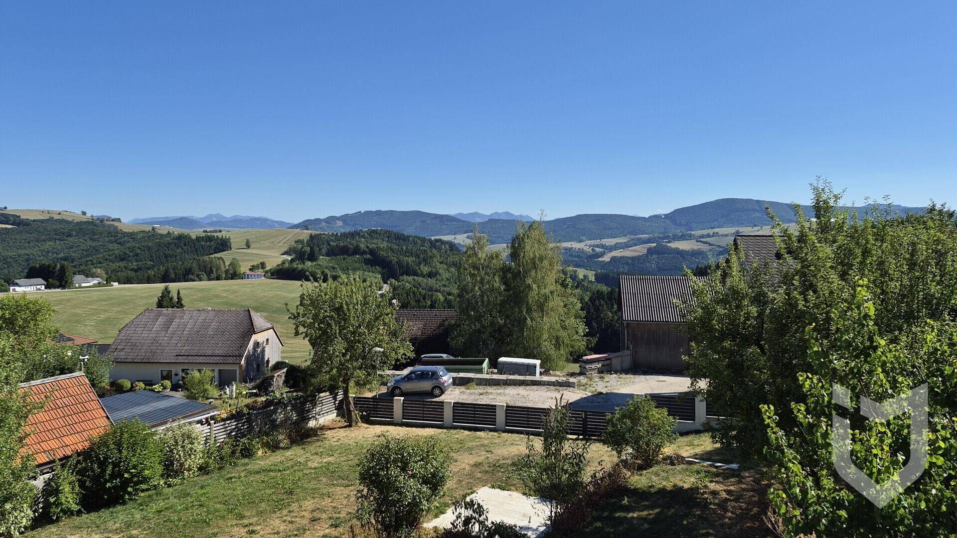 Weitläufiger Blick über die grüne Hügellandschaft mit Bergen im Hintergrund unter blauem Himmel.
