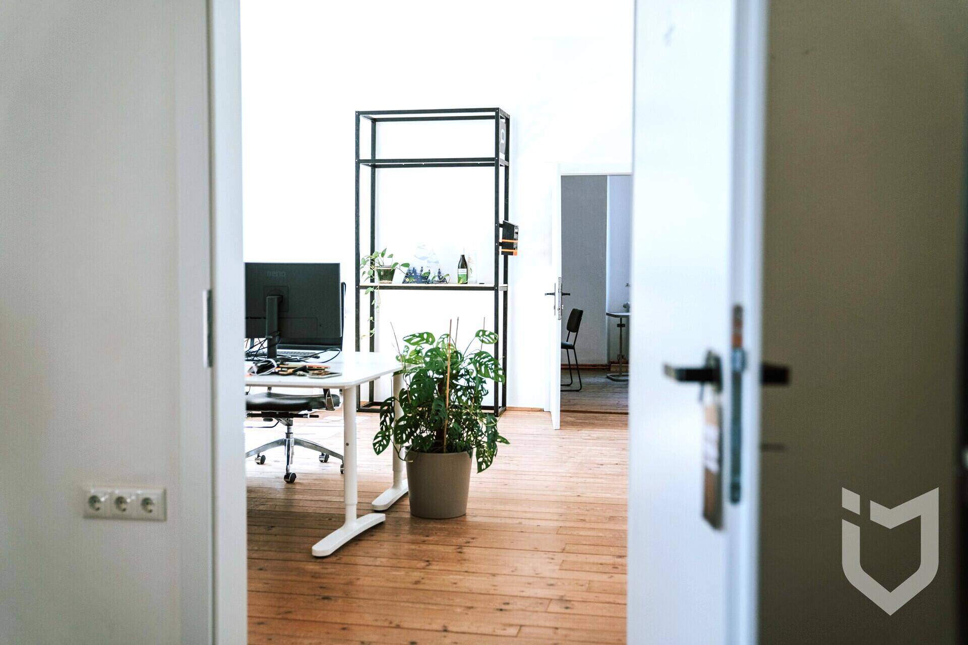 Heller Büroraum mit Holzdielenboden, modernem Schreibtisch und Pflanzen, gesehen durch eine offene Tür.