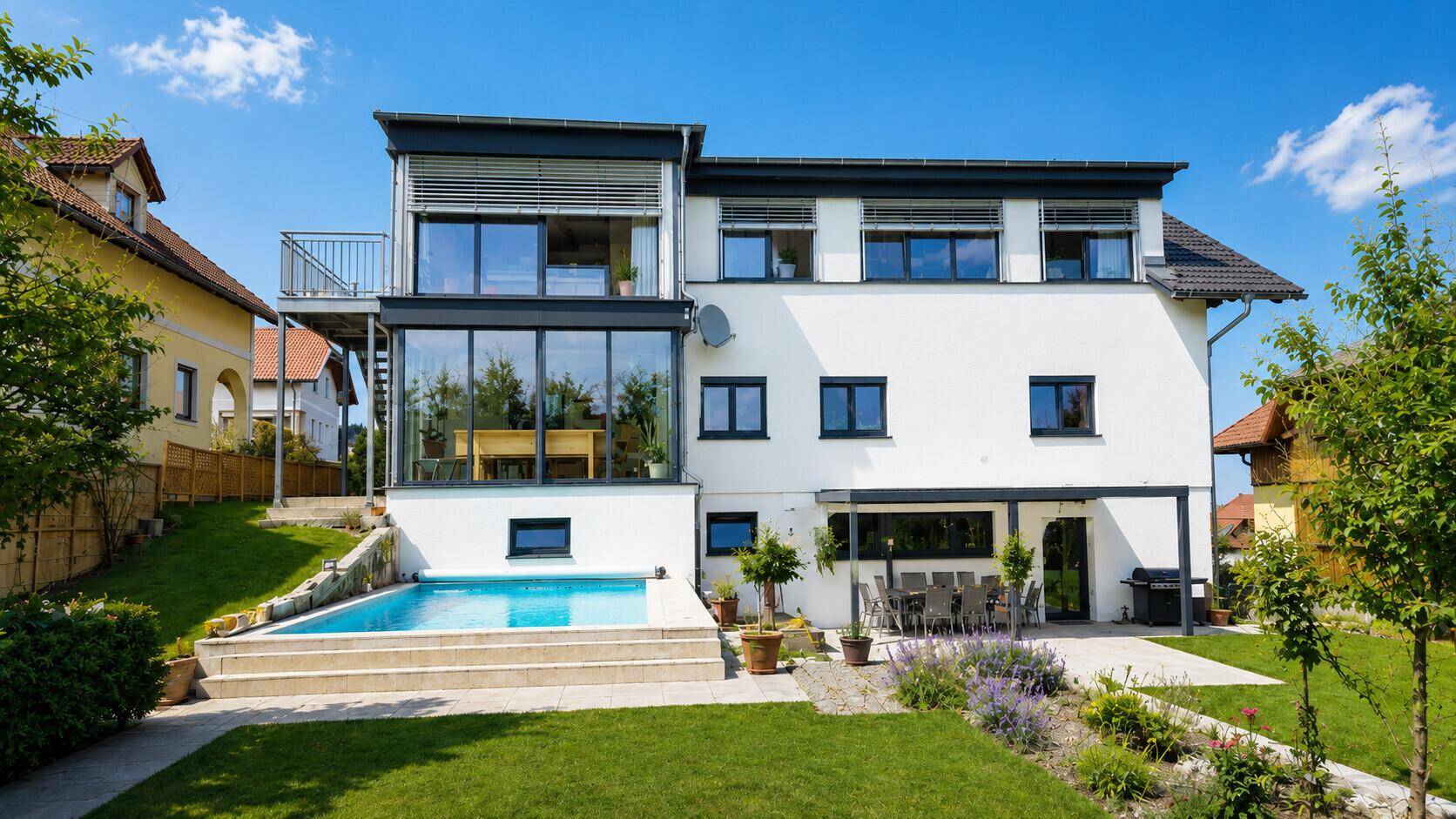 Modernes Zweifamilienhaus mit Pool und Garten, umgeben von grüner Landschaft unter blauem Himmel.