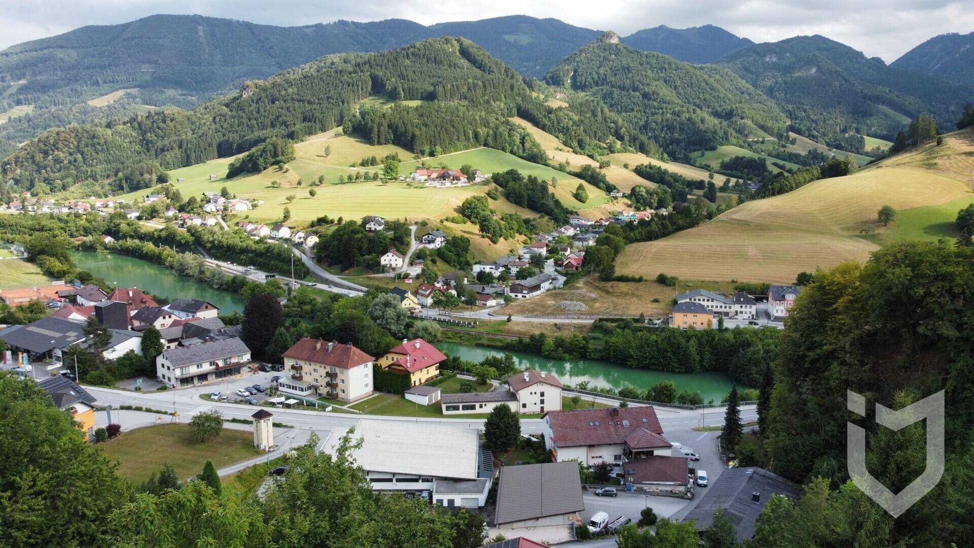 Panoramablick auf die malerische Ortschaft in einem Tal mit Fluss und bewaldeten Bergen.