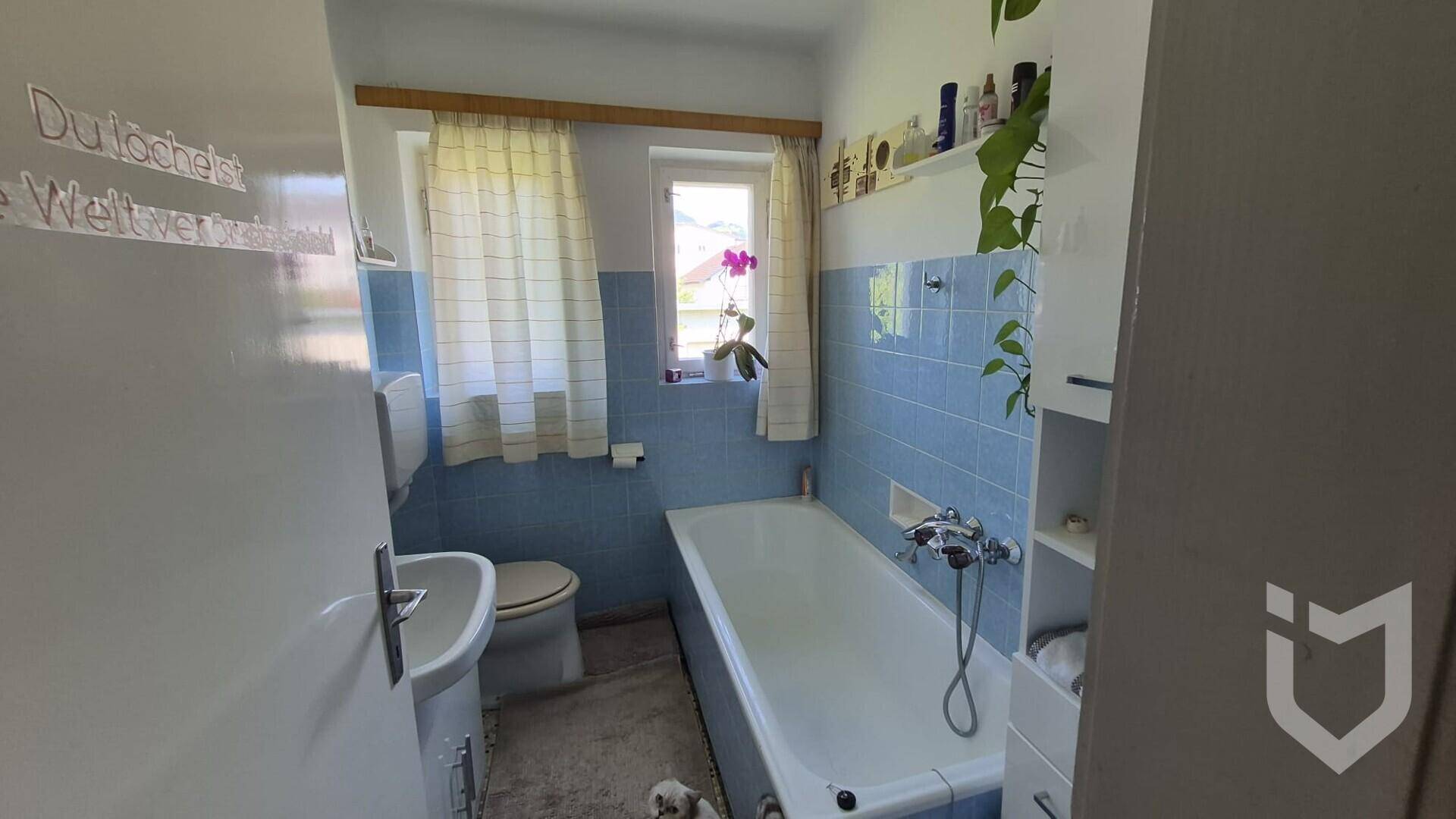 Badezimmer mit blauen Fliesen, Badewanne, WC und Fenster, im Stil der 80er Jahre.