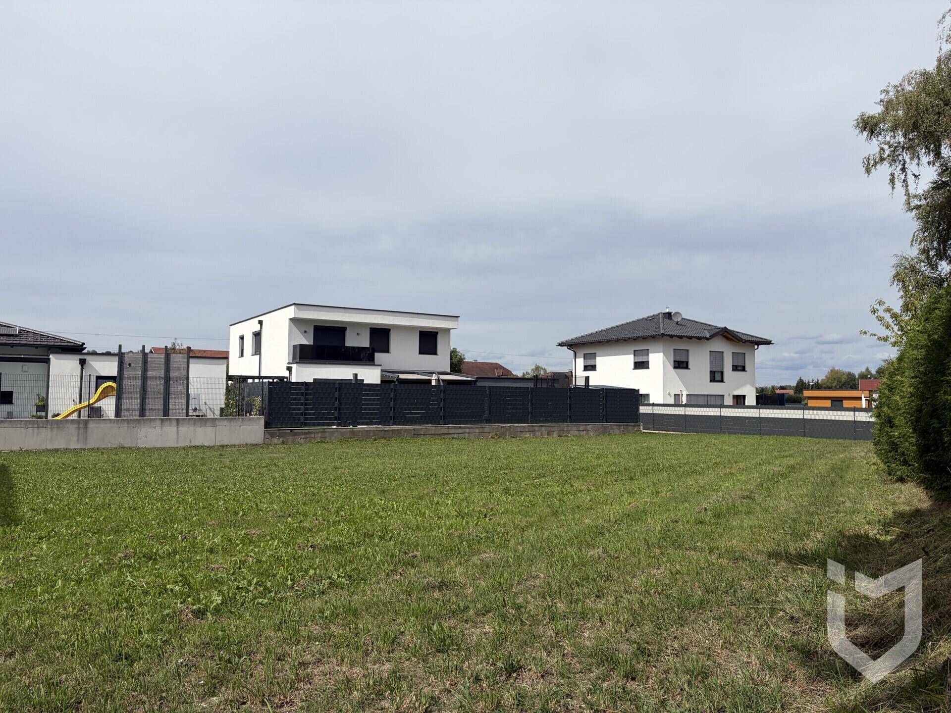 Offenes Baugrundstück mit Blick auf moderne Einfamilienhäuser und einen kleinen Spielbereich im Hintergrund.
