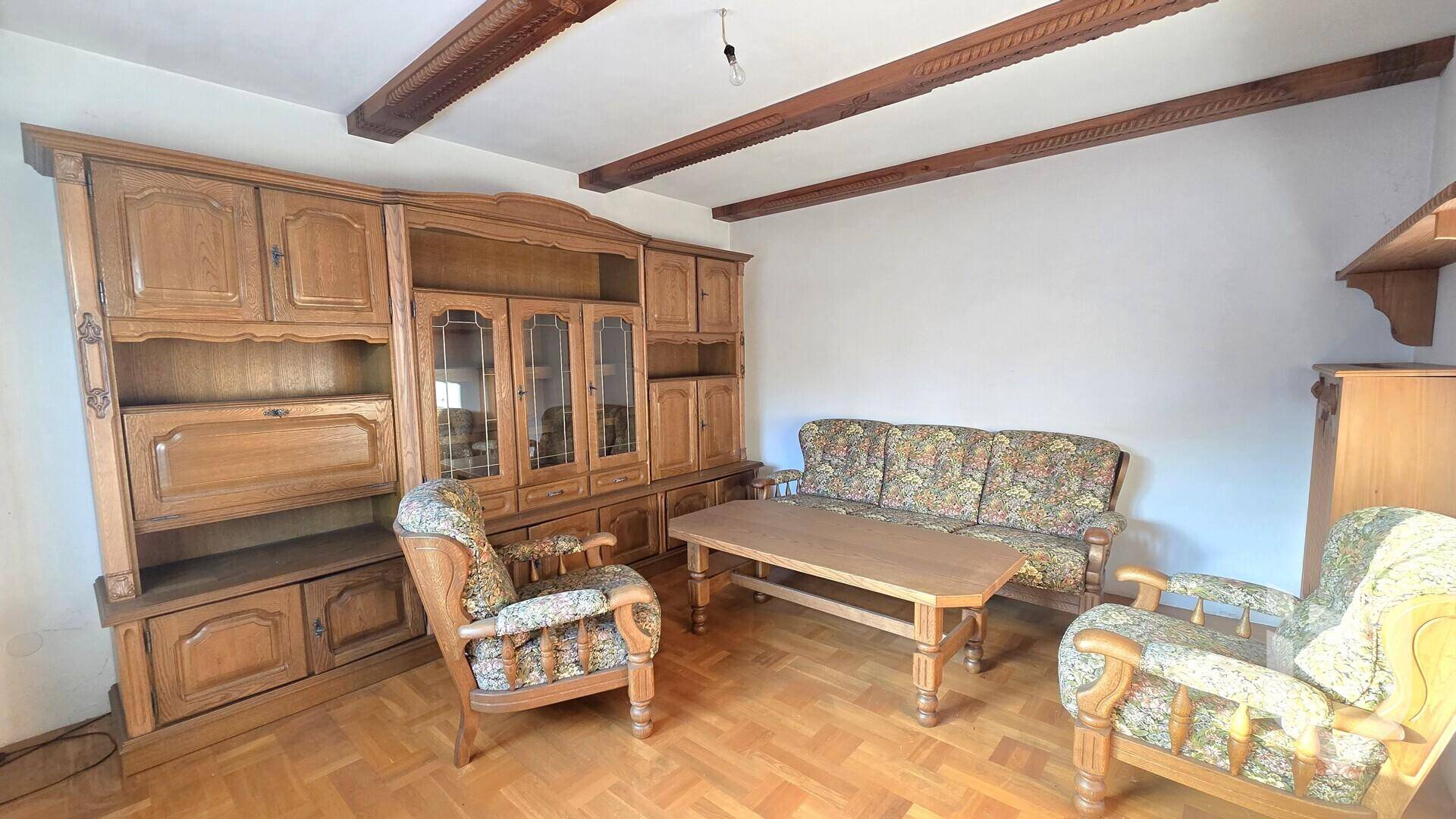 Wohnzimmer mit rustikalen Holzmöbeln, gemustertem Sofa und sichtbaren Deckenbalken, das eine Renovierung benötigt.