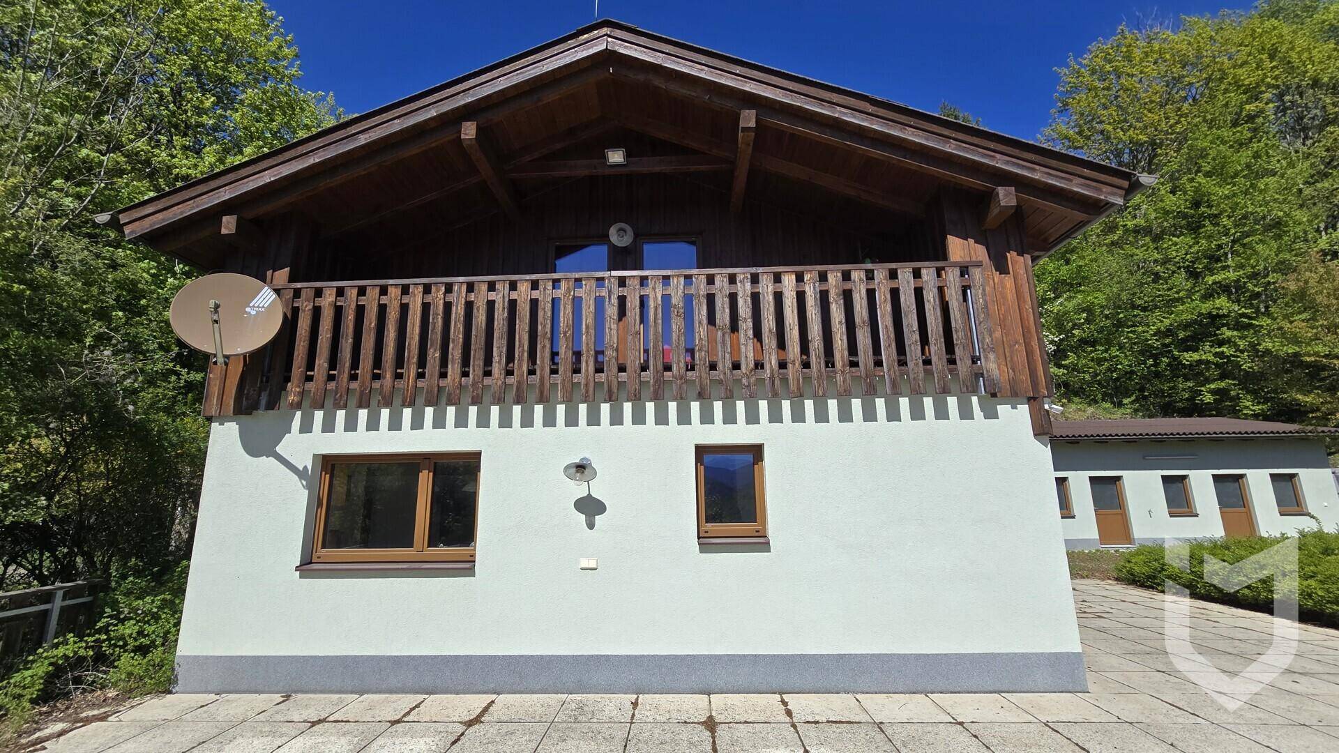 Fassade des Hauses mit Holzbalkon im Obergeschoss und einem Nebengebäude im Hintergrund der Natur.