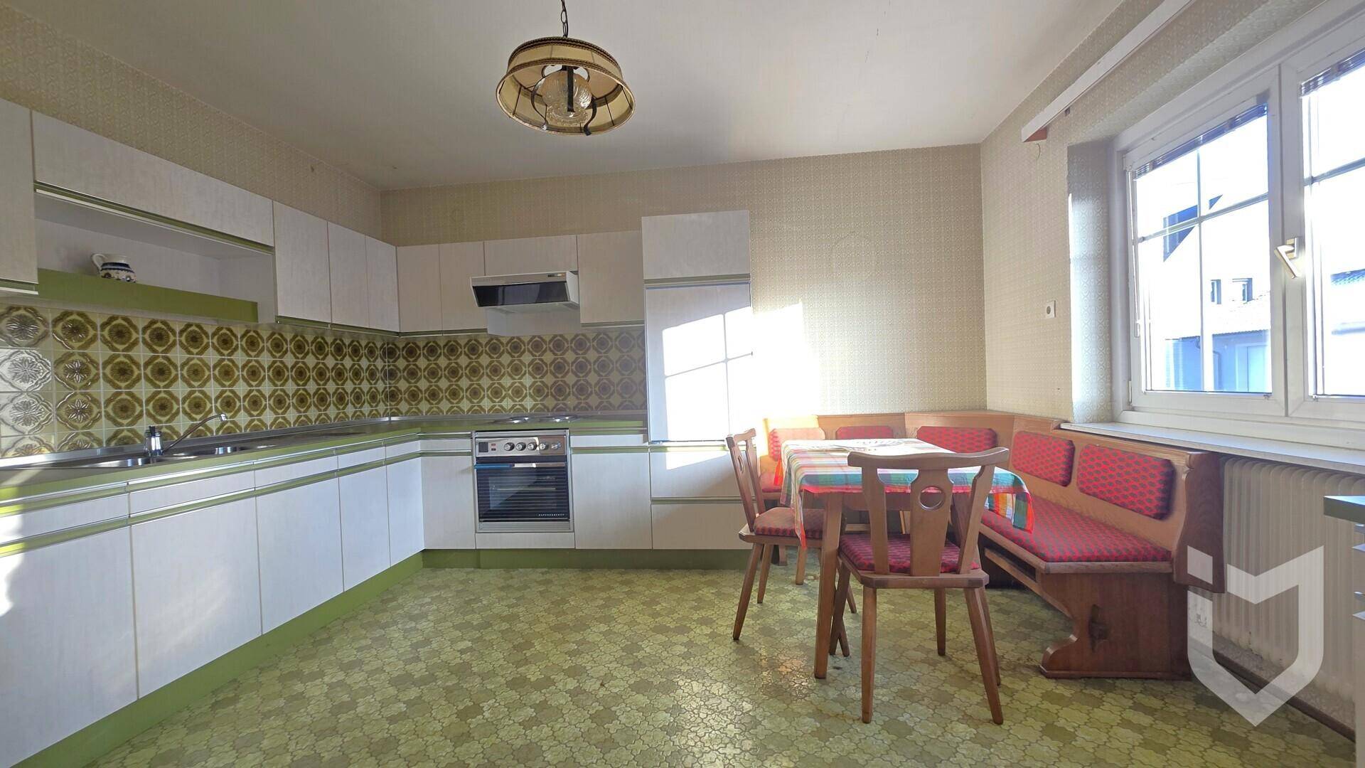 Geräumige Wohnküche mit weißen Einbauschränken, grün gemusterten Fliesen und einer Essecke am Fenster.