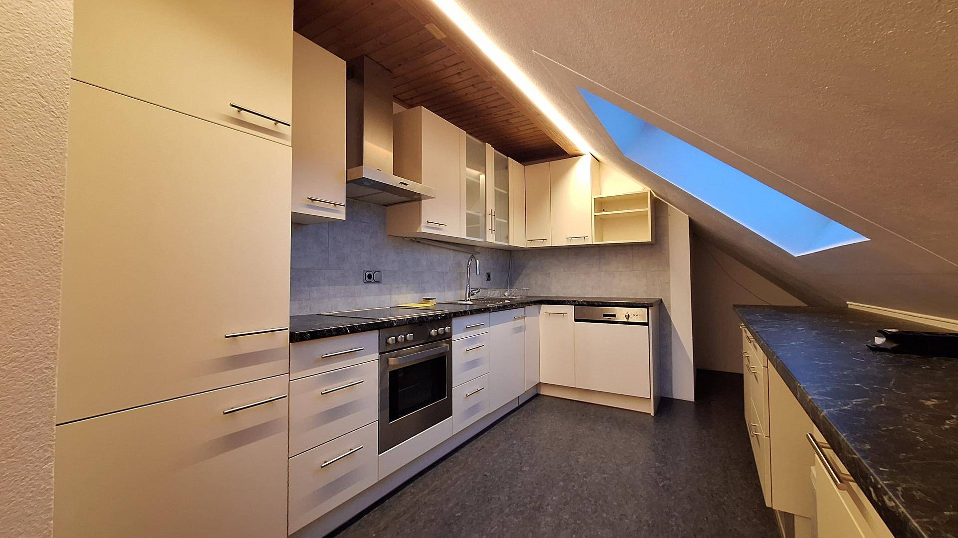 Moderne Einbauküche im Dachgeschoss mit hellen Schränken, dunkler Arbeitsplatte und Dachfenster.