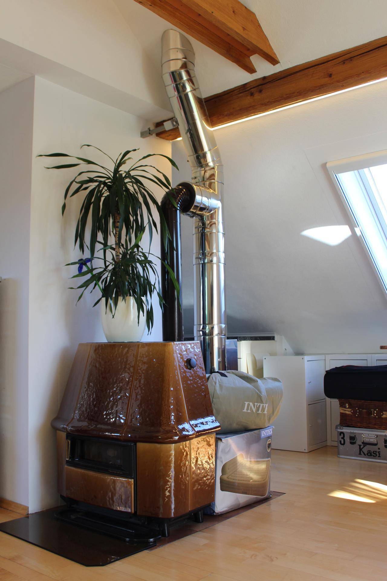 Rustikaler Kachelofen mit metallenem Ofenrohr und Holzbalken in einem hellen Dachgeschossraum.