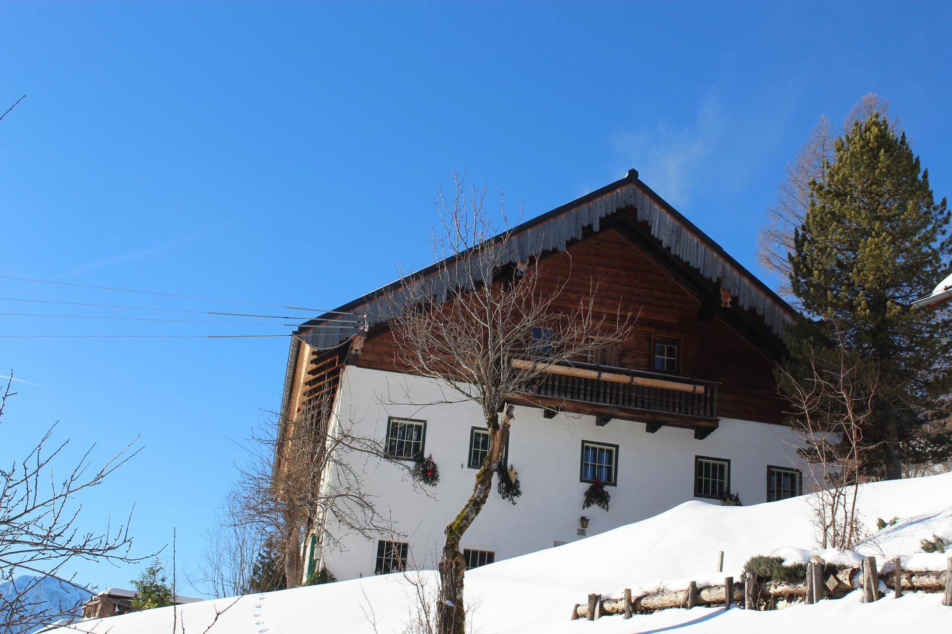 Traditionelles Bauernhaus im Winter, umgeben von Schnee und kahlen Bäumen unter blauem Himmel.