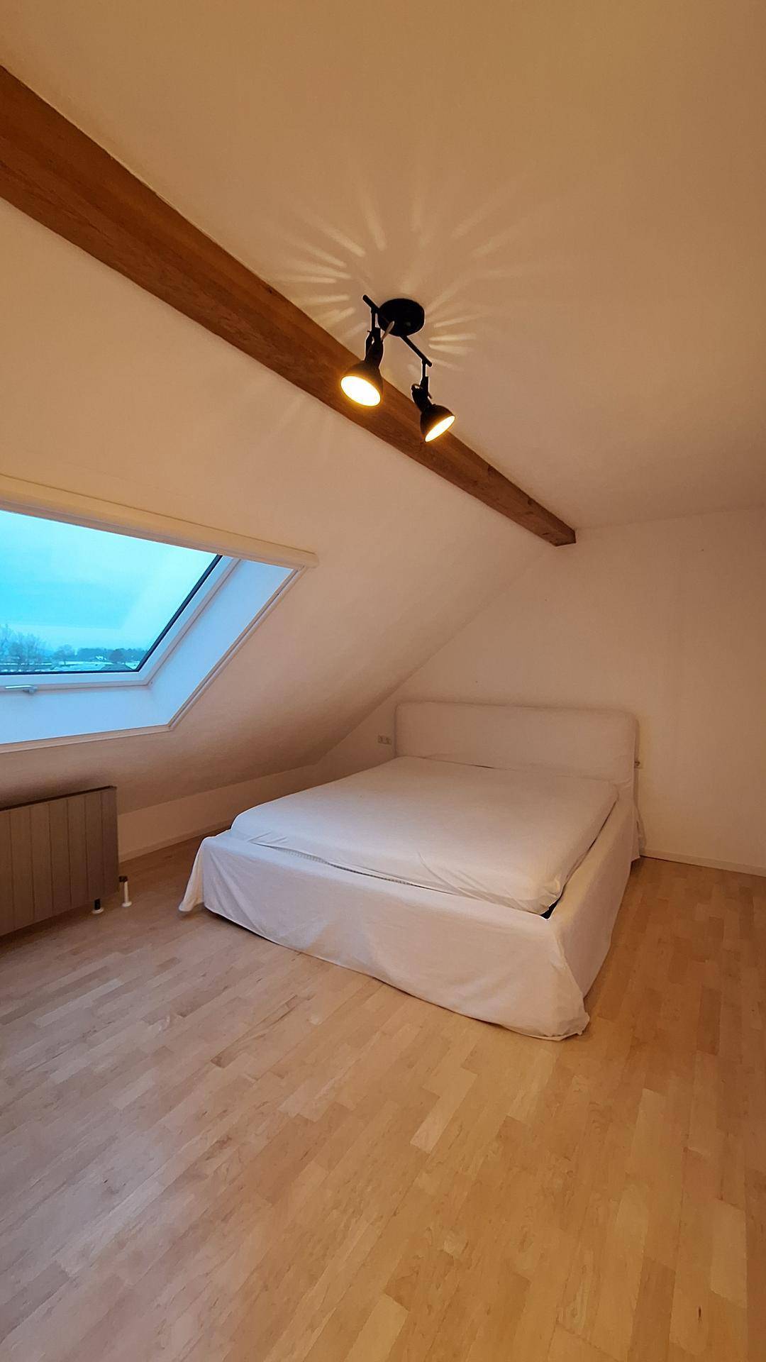 Helles Schlafzimmer im Dachgeschoss mit Holzbalken, Dachfenster und hellem Holzfußboden.