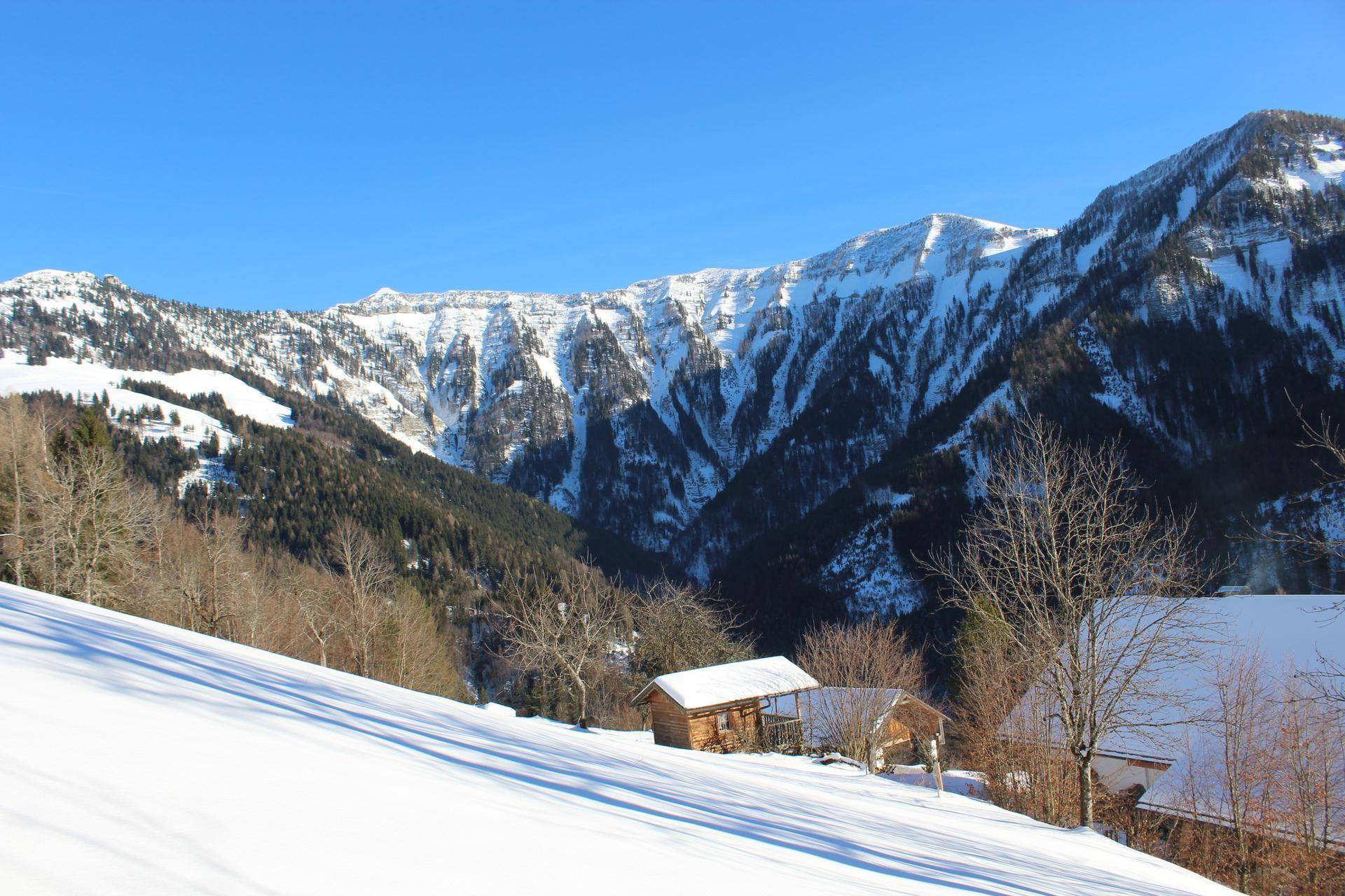 Weitläufige schneebedeckte Berglandschaft mit kleinen Hütten und Wäldern unter blauem Himmel.