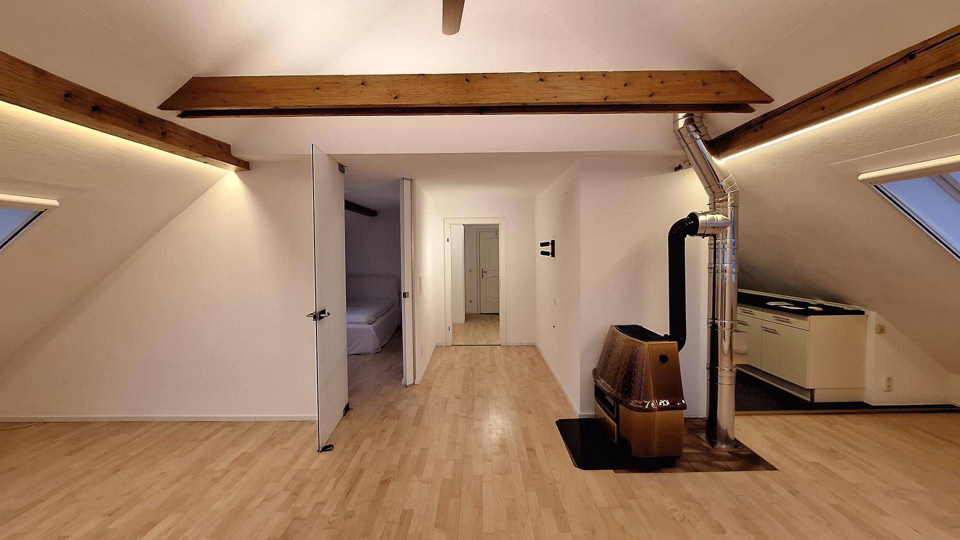 Offener Dachgeschoss-Wohnbereich mit Kaminofen, Holzbalken und Zugang zu weiteren Räumen.