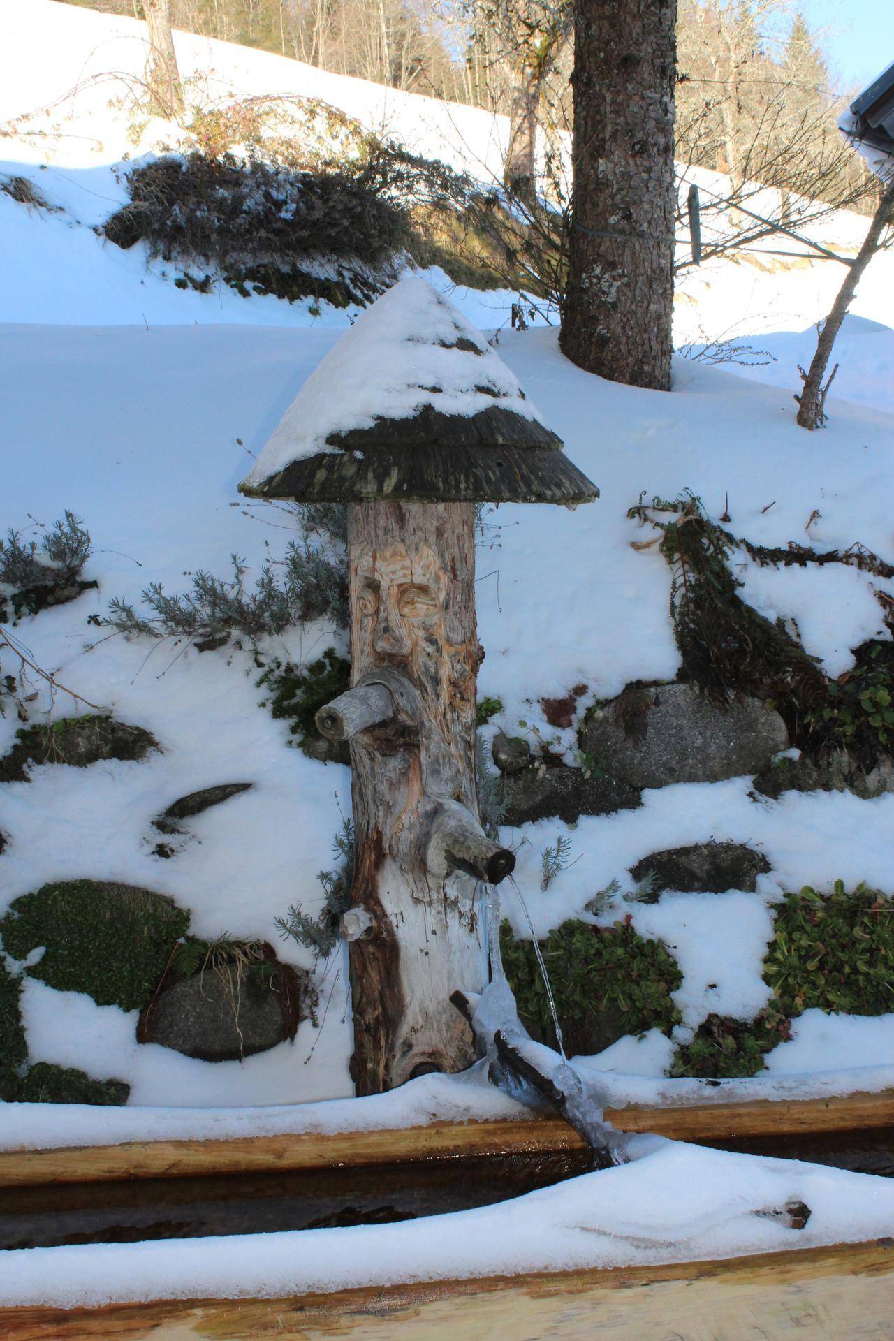 Rustikaler Holzbrunnen im verschneiten Garten, umgeben von Schnee und Bäumen.