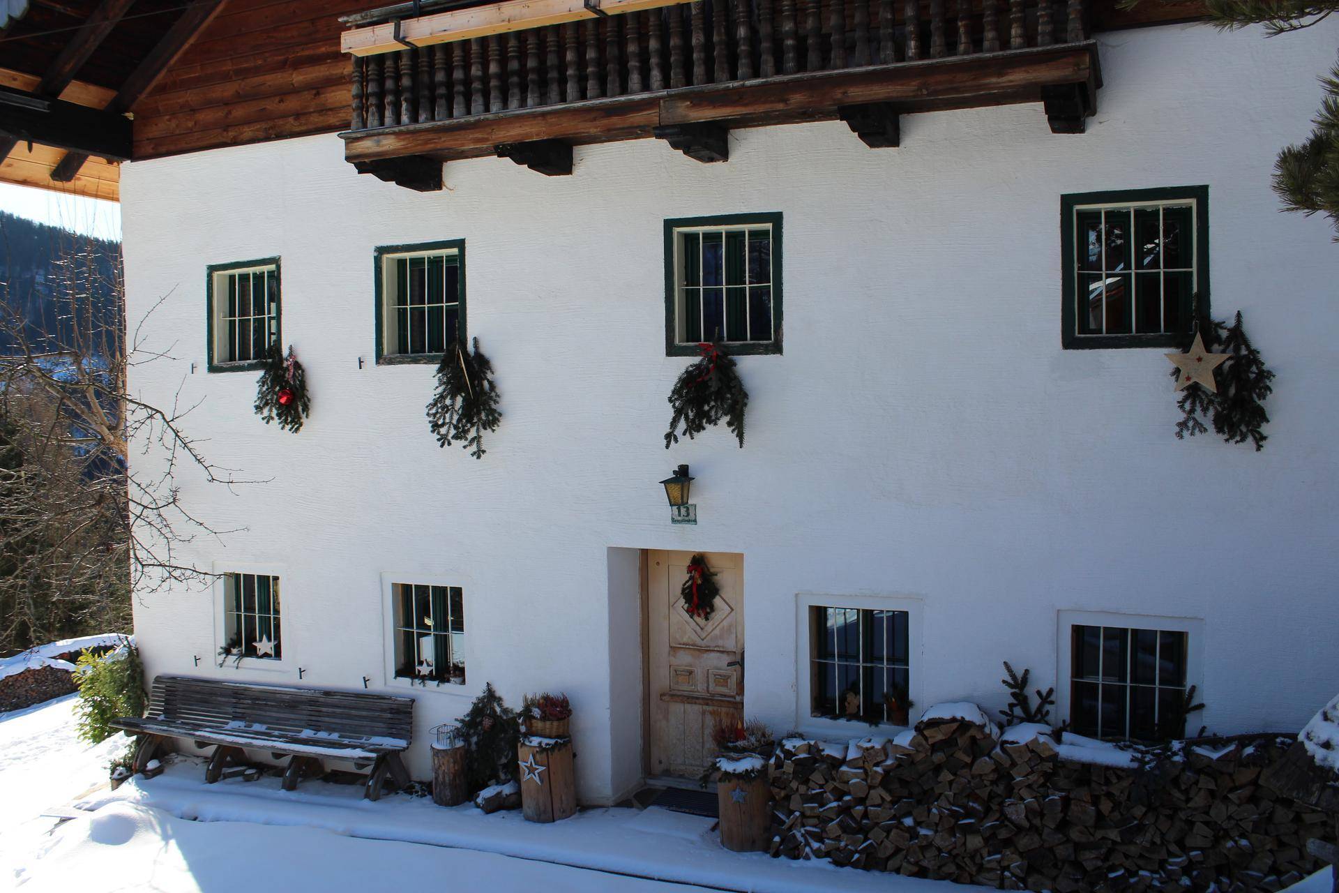 Traditionelles Bauernhaus im Winter mit verschneitem Vorplatz, Holzstapel und festlicher Dekoration an den Fenstern.