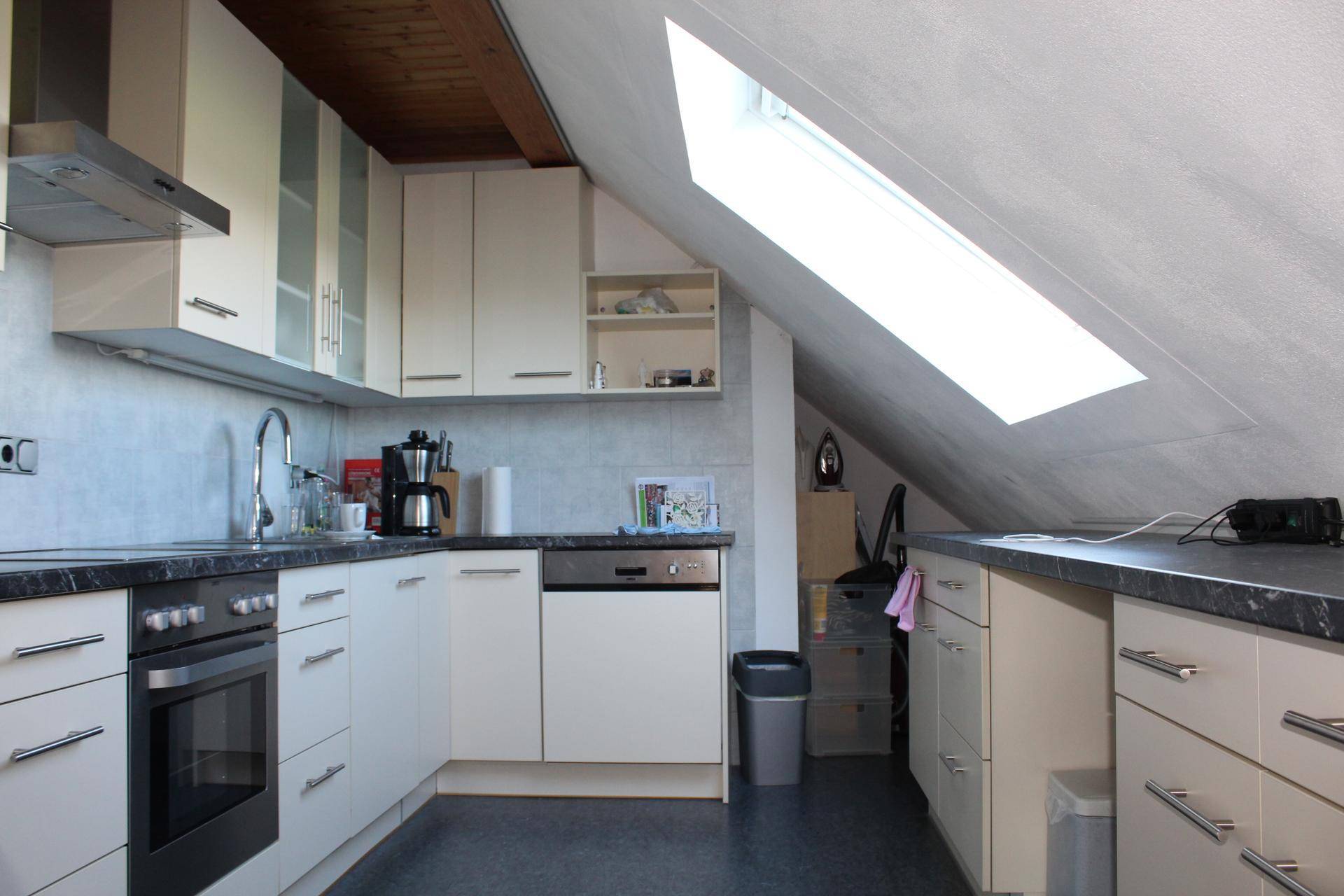 Helle Küche im Dachgeschoss mit Schrägdachfenster, weißen Schränken und dunkler Arbeitsplatte.