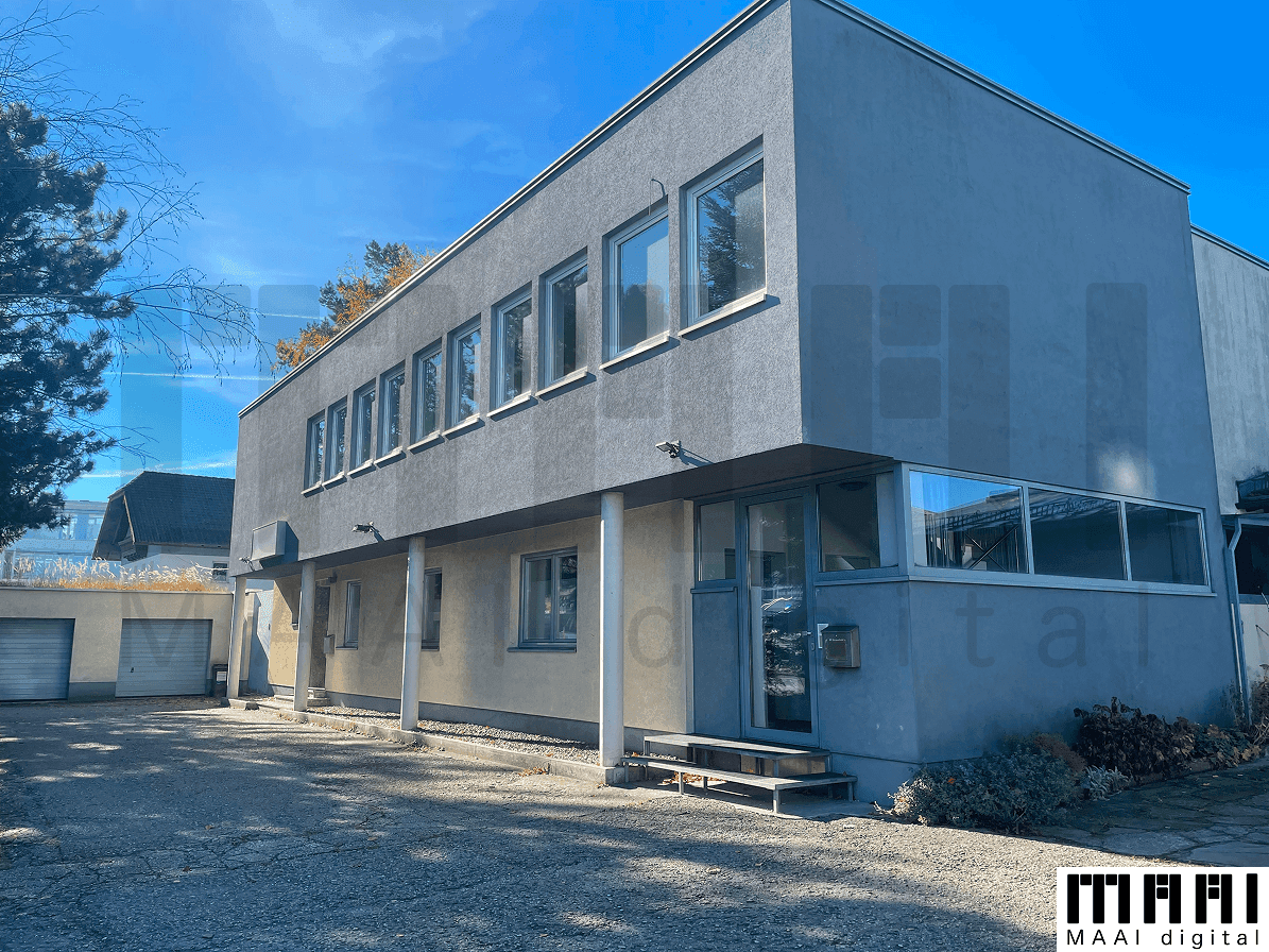 Modernes Gebäude mit grauer Fassade, großen Fenstern und einem Flachdach, umgeben von einer Zufahrt.