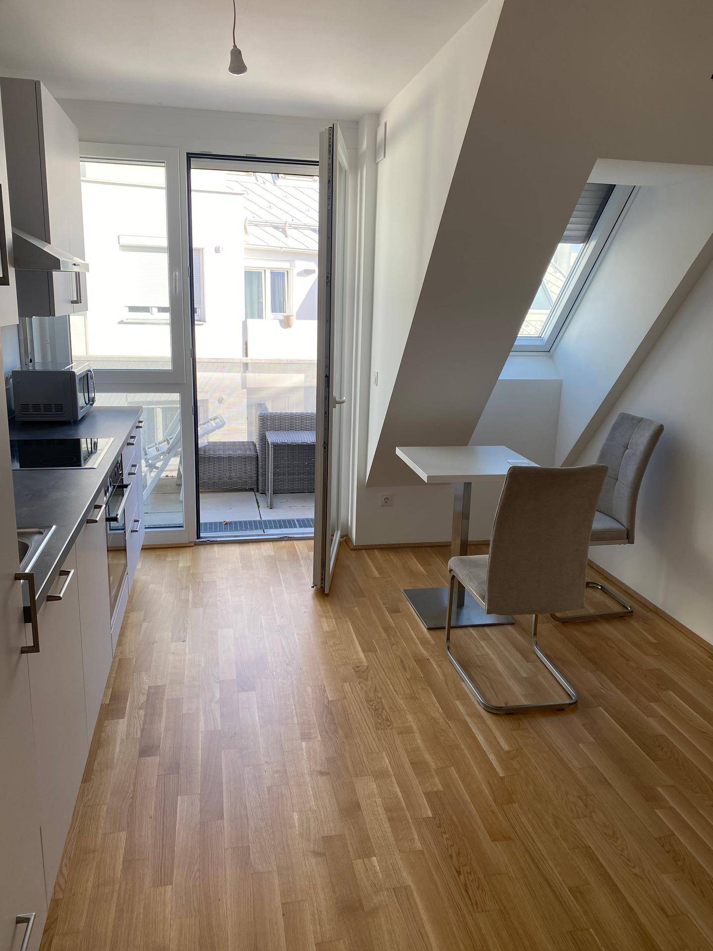 Helle Küche mit Holzboden, kleinem Essbereich und Zugang zum Balkon unter Dachschrägen.