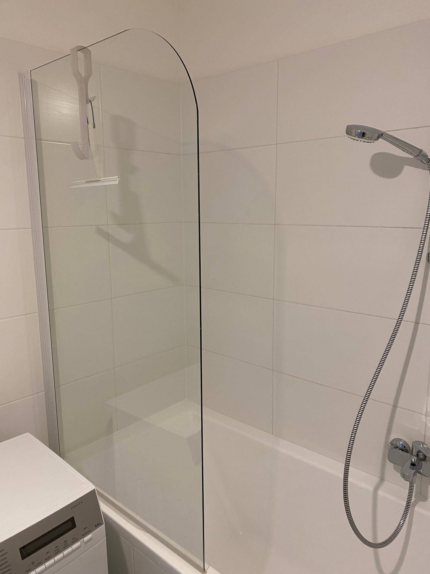 Detailansicht der Badewanne mit Glasabtrennung und Duschkopf in einem hell gefliesten Bad.