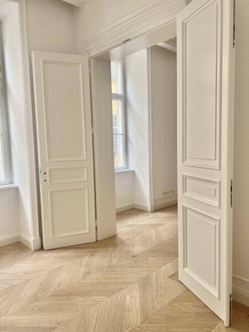 Heller Raum mit elegantem Fischgrätparkett, hohen weißen Türen und viel Tageslicht durch große Fenster.