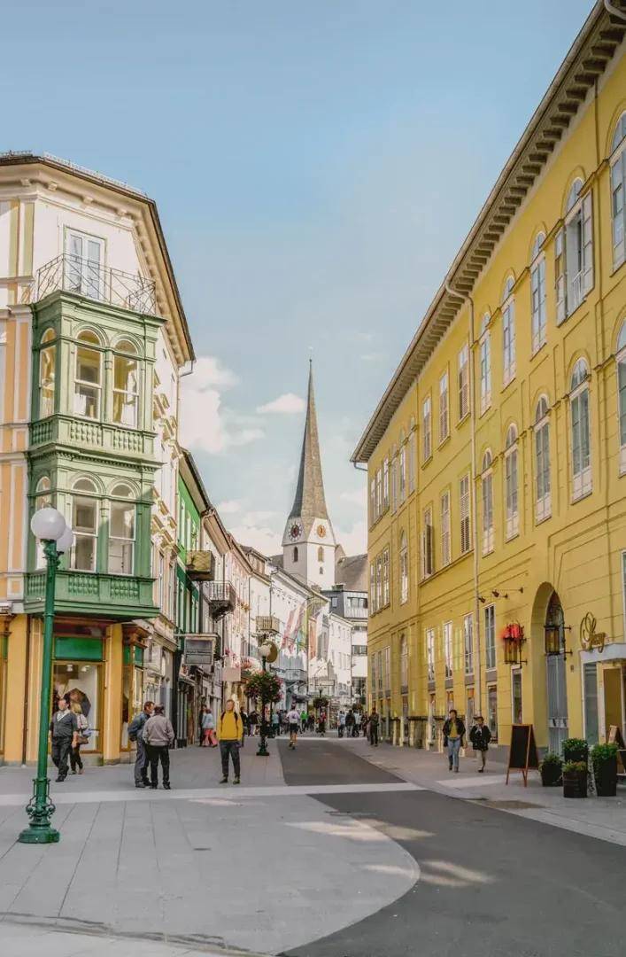 Belebte historische Gasse mit charmanten Gebäudefassaden, Erkern und einem markanten Kirchturm unter blauem Himmel.