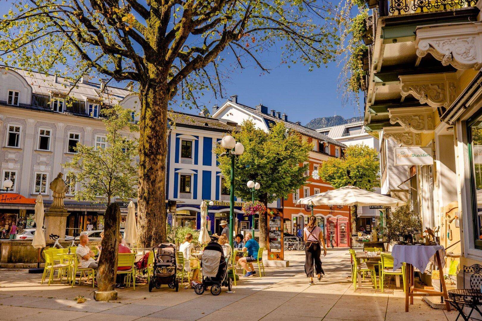 Belebter Stadtplatz mit Straßencafés, bunten Gebäudefassaden und einem großen schattenspendenden Baum.