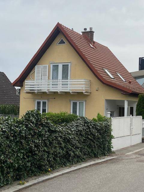 Gelbes Einfamilienhaus mit rotem Satteldach, weißem Balkon und grüner Hecke im Vorgarten.