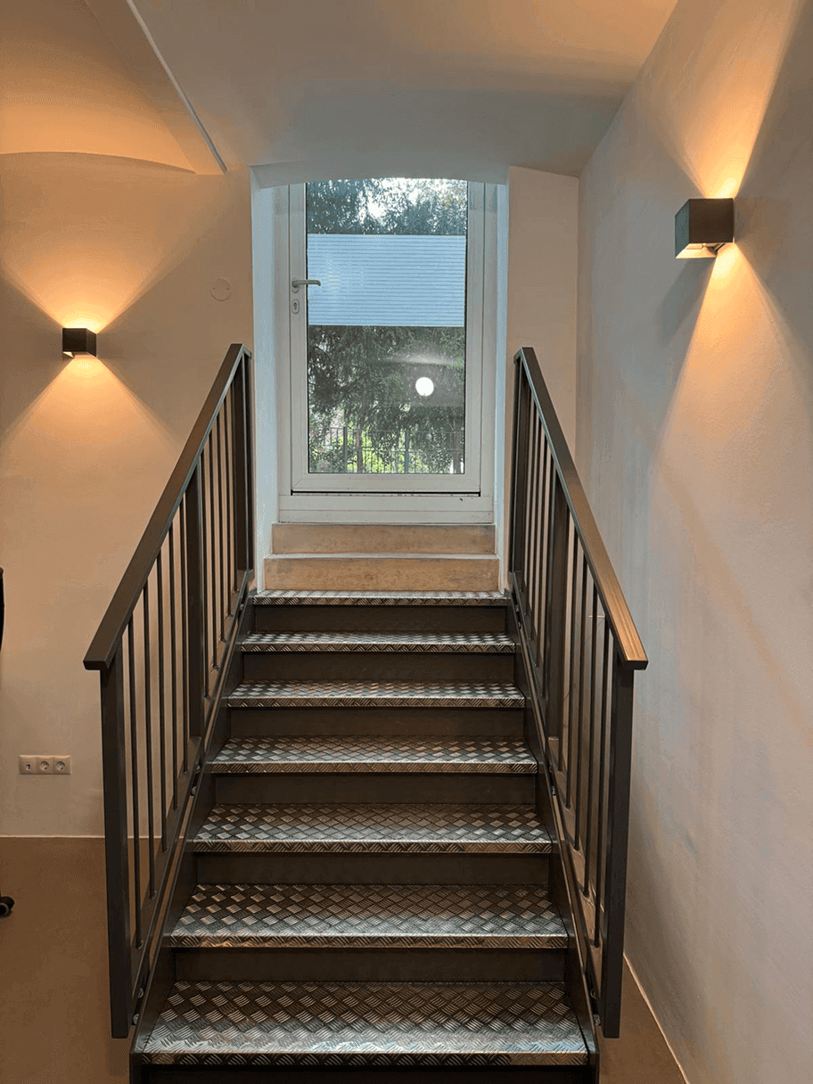 Industrielle Metalltreppe mit Riffelblechstufen, die zu einer Tür mit Fenster und Wandleuchten führt.