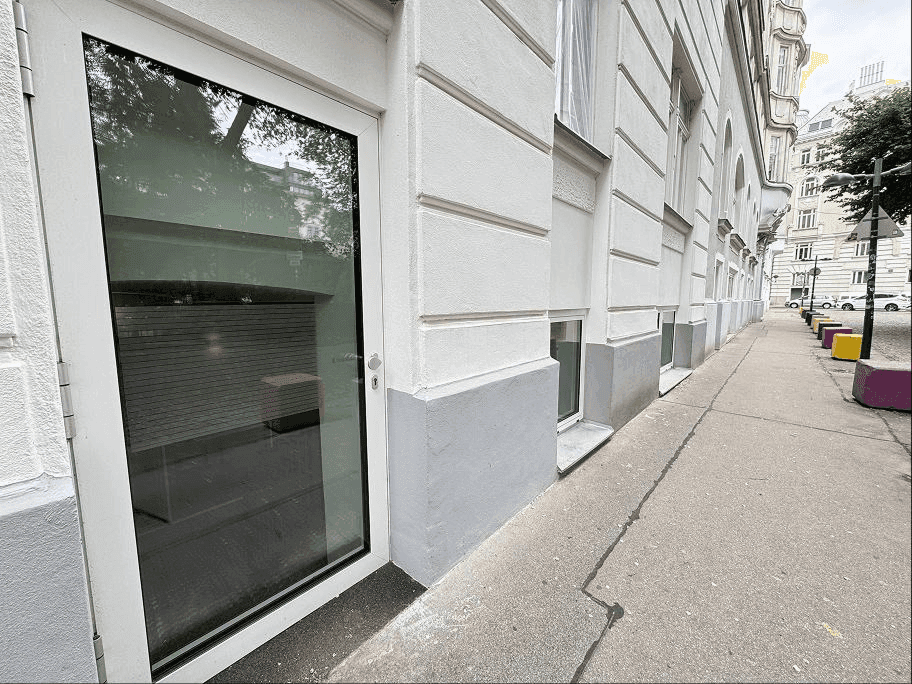 Straßenansicht einer Gebäudefassade mit großer Glastür und Fenstern, Bürgersteig und Straße im Hintergrund.