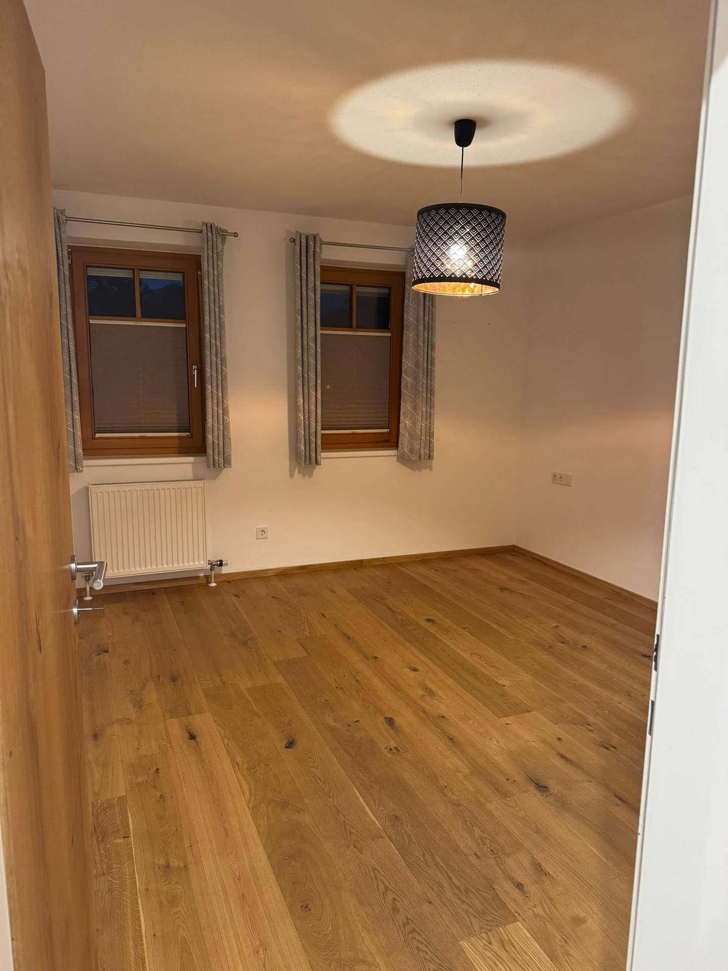 Leeres Zimmer mit Holzboden, zwei Fenstern und einer Deckenleuchte, bereit zur individuellen Gestaltung.