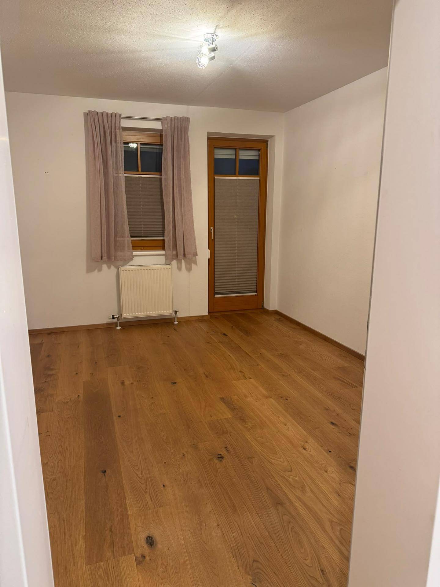 Helles Zimmer mit Holzboden, Fenster und einer Tür, die zu einem Balkon oder Garten führen könnte.