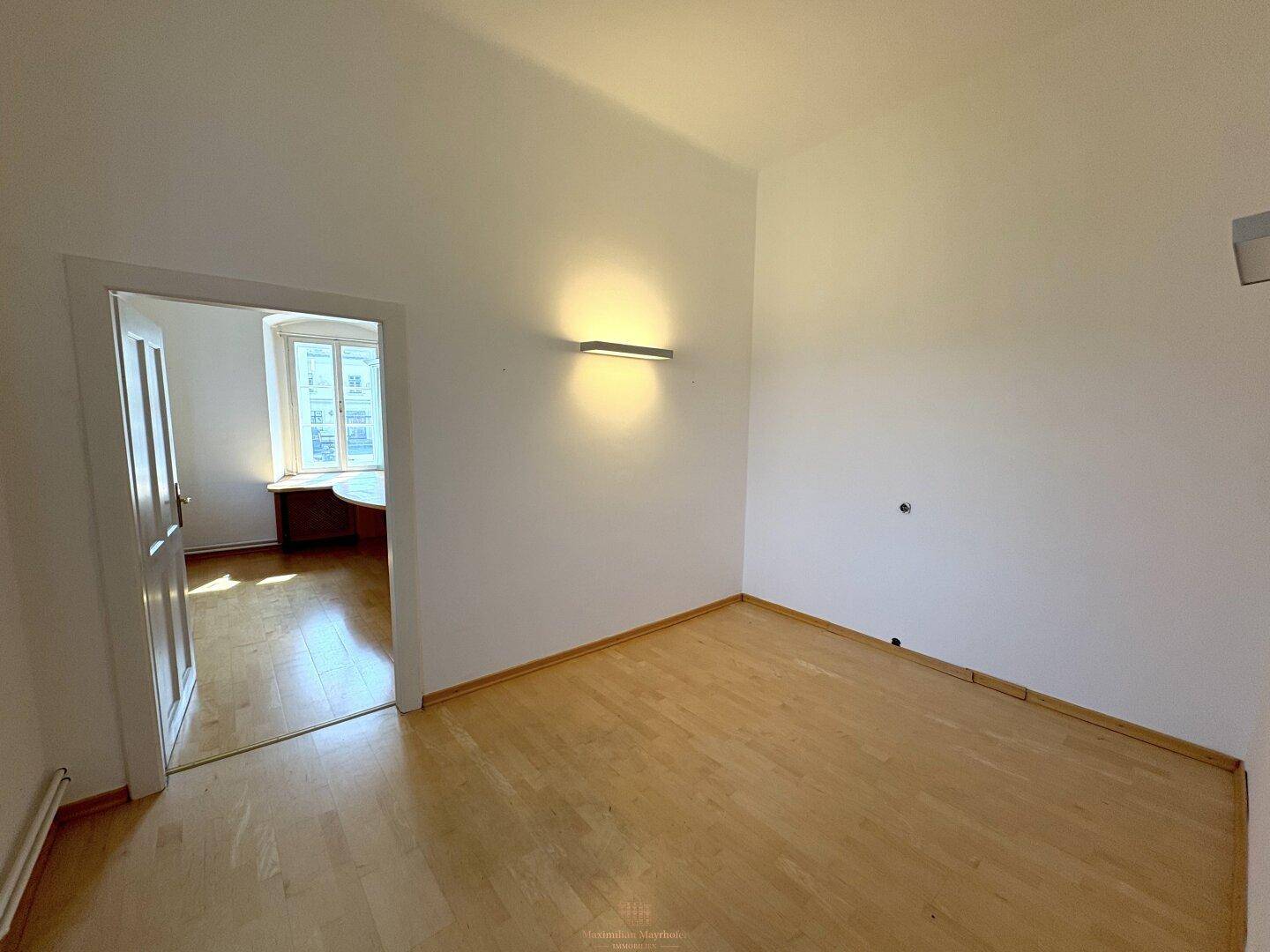 Leerer Raum mit Holzboden und weiß gestrichenen Wänden, der zu einem weiteren Zimmer führt.