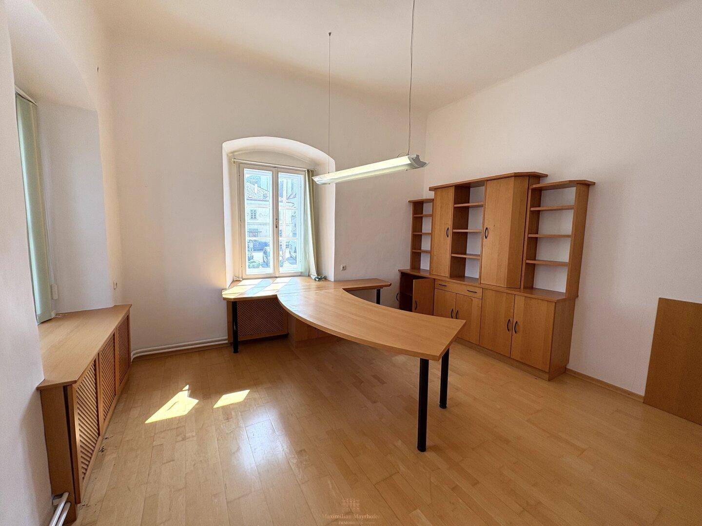 Geräumiges Büro mit Holzboden, Schreibtisch und Schränken, beleuchtet durch ein großes Fenster.