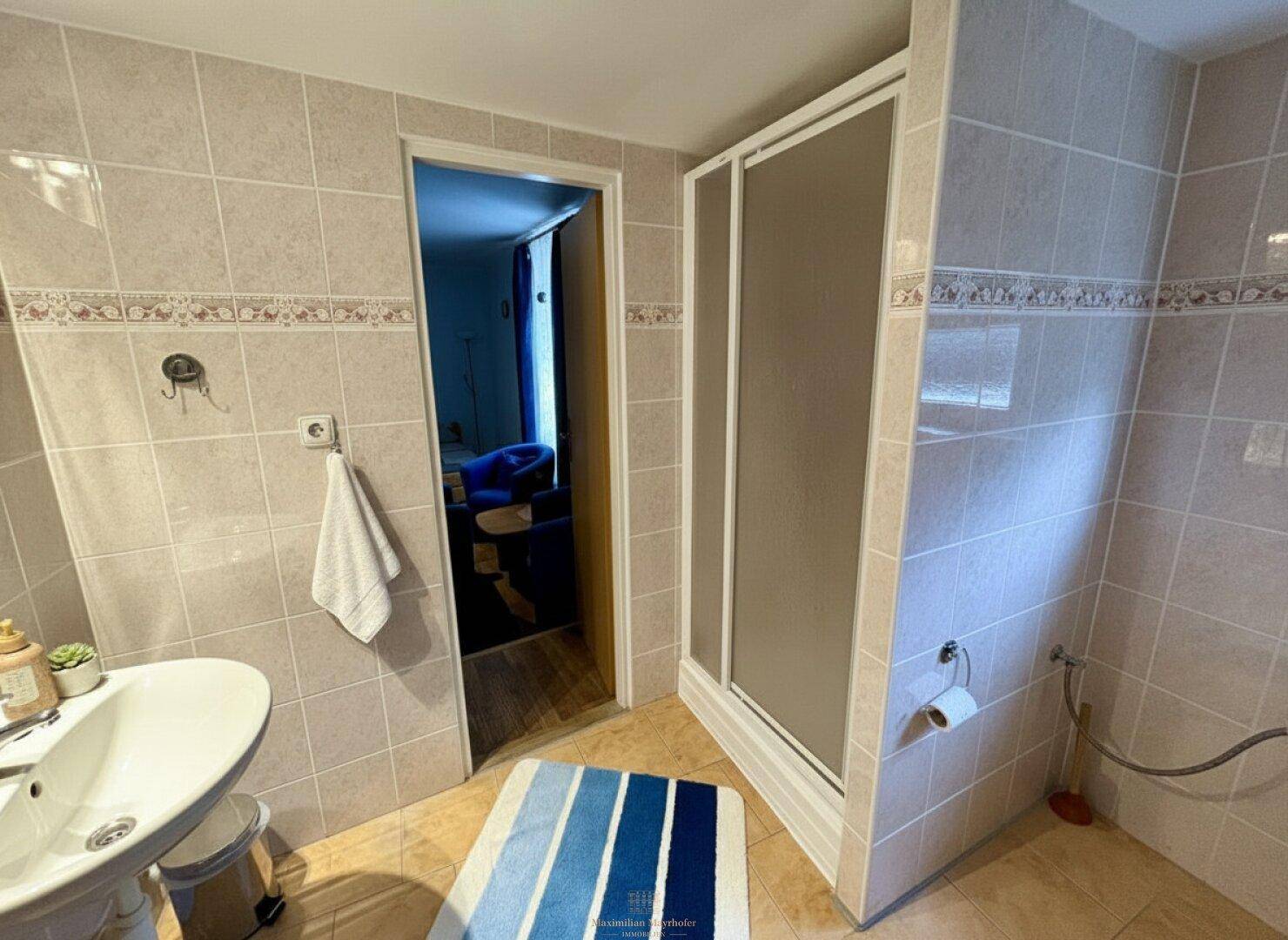 Gefliestes Badezimmer mit Duschkabine, Waschbecken und einem blauen Badteppich.