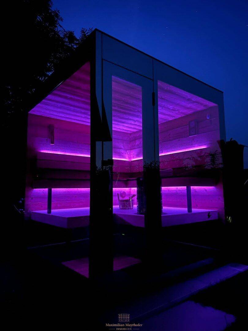 Moderne Saunakabine im Garten bei Nacht, beleuchtet mit lila LED-Lichtern.