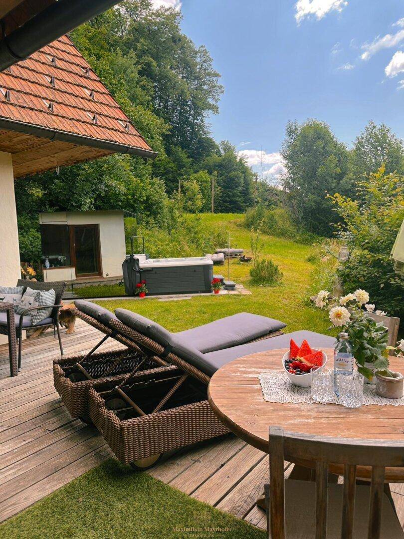 Sonnige Holzterrasse mit Esstisch, Liegestühlen und Blick auf den Garten und Whirlpool.