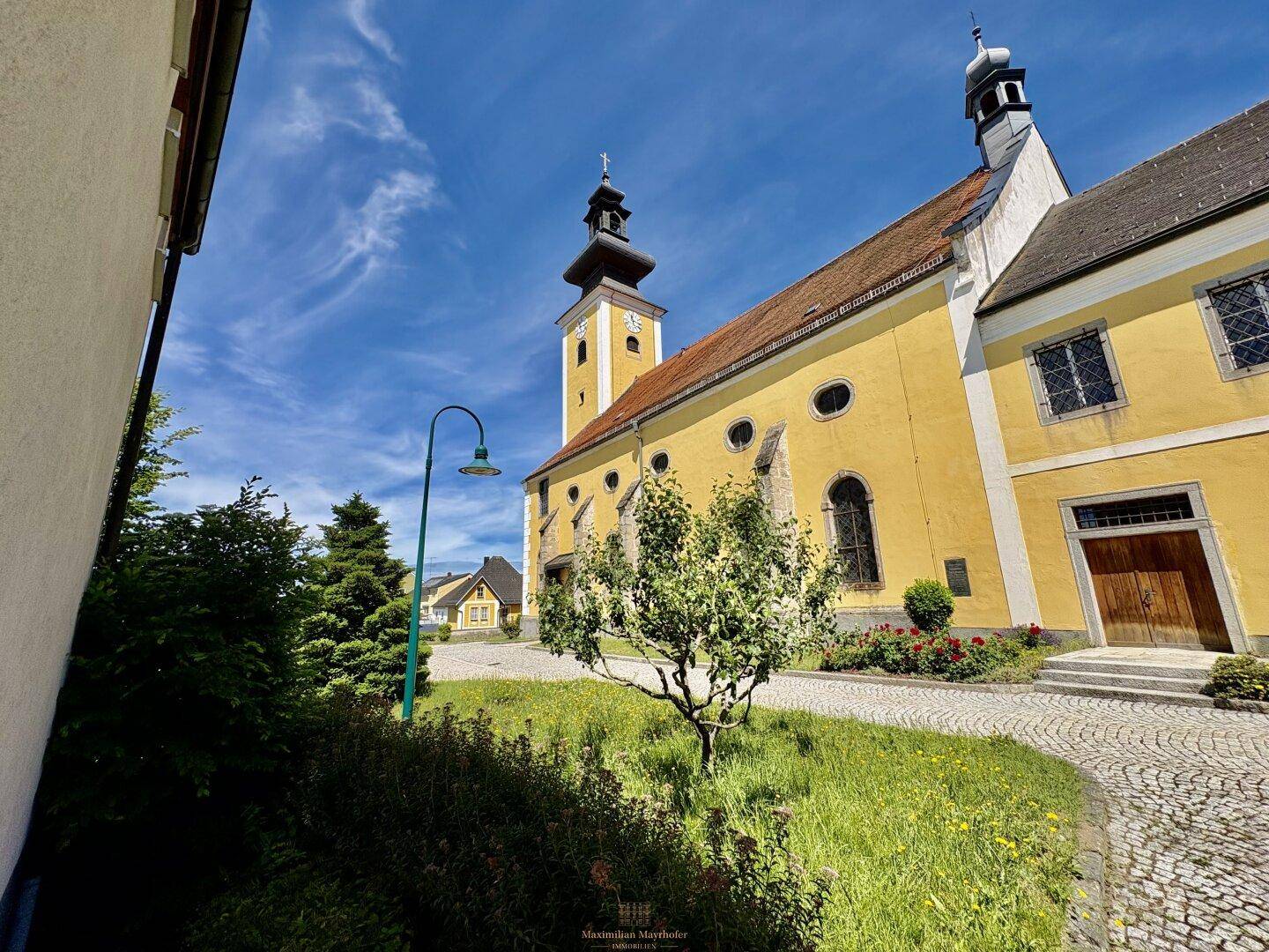 Historisches Kirchengebäude mit gelber Fassade und gepflegtem Vorplatz unter blauem Himmel.