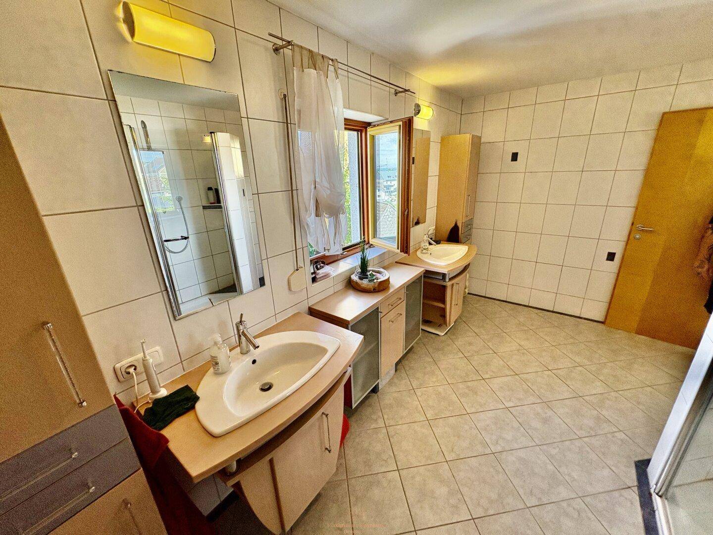 Badezimmer mit zwei Waschbecken, großem Spiegel und Fenster, das viel Licht spendet.
