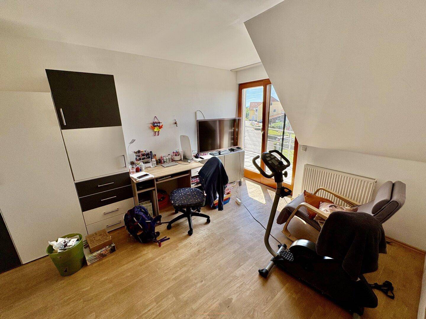Schlafzimmer mit Arbeitsbereich, Fitnessgerät und Zugang zu einem kleinen Balkon.