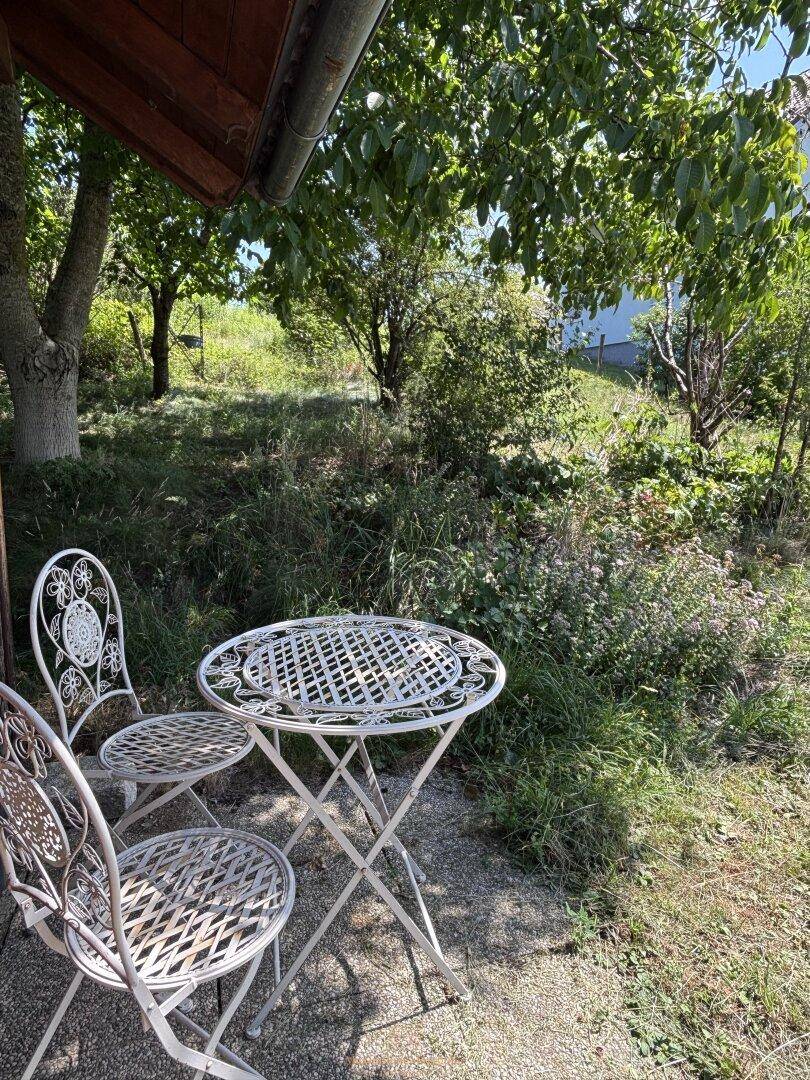Gemütlicher Sitzbereich im Freien mit weißem Metalltisch und Stühlen unter schattenspendenden Bäumen.