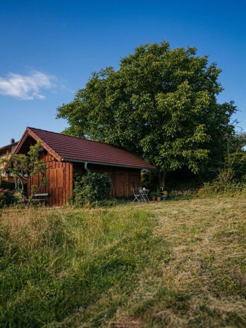 Rustikales Holzhaus mit kleiner Terrasse und Gartenmöbeln, eingebettet in eine grüne Landschaft.