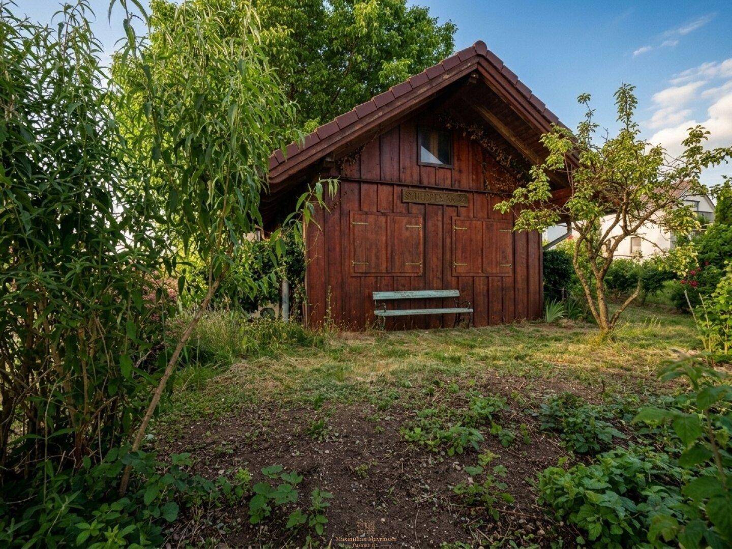 Hölzernes Gartenhaus mit kleiner Bank und Fensterläden, umgeben von Bäumen und einer Wiese.