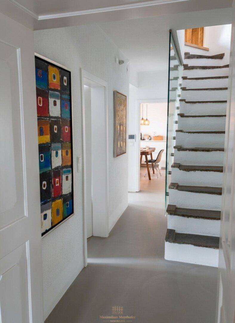 Heller Flur mit moderner Kunst, Treppe mit Glasgeländer und Blick in den Essbereich.