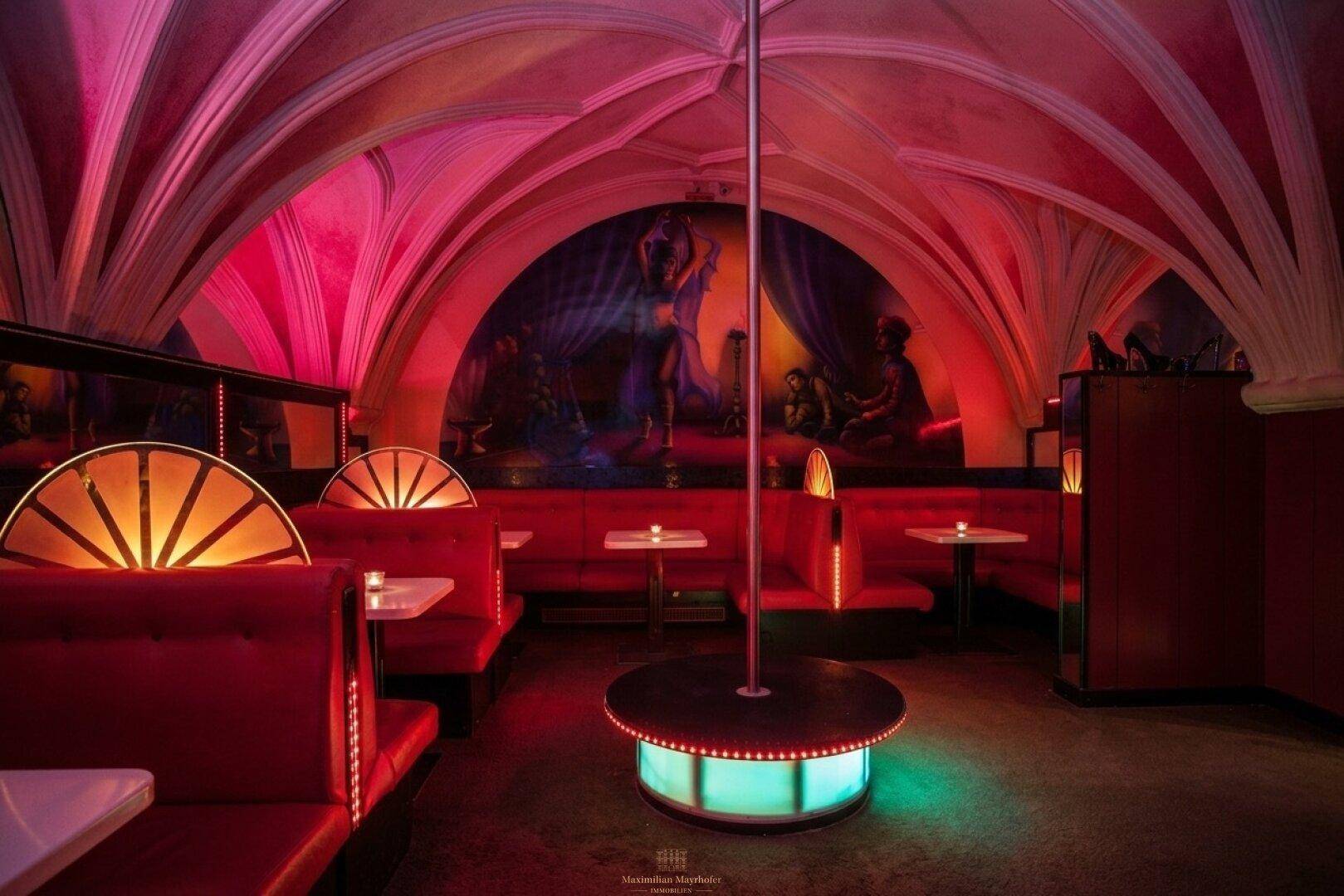 Innenbereich einer Bar mit roter Beleuchtung, Gewölbedecken, roten Sitzbänken und einer Tanzstange.