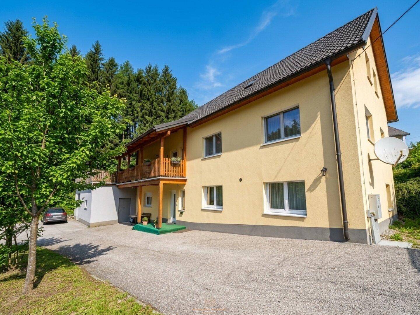 Ansicht des gelben Hauses mit Holzbalkon und großem Vorplatz, umgeben von Bäumen.