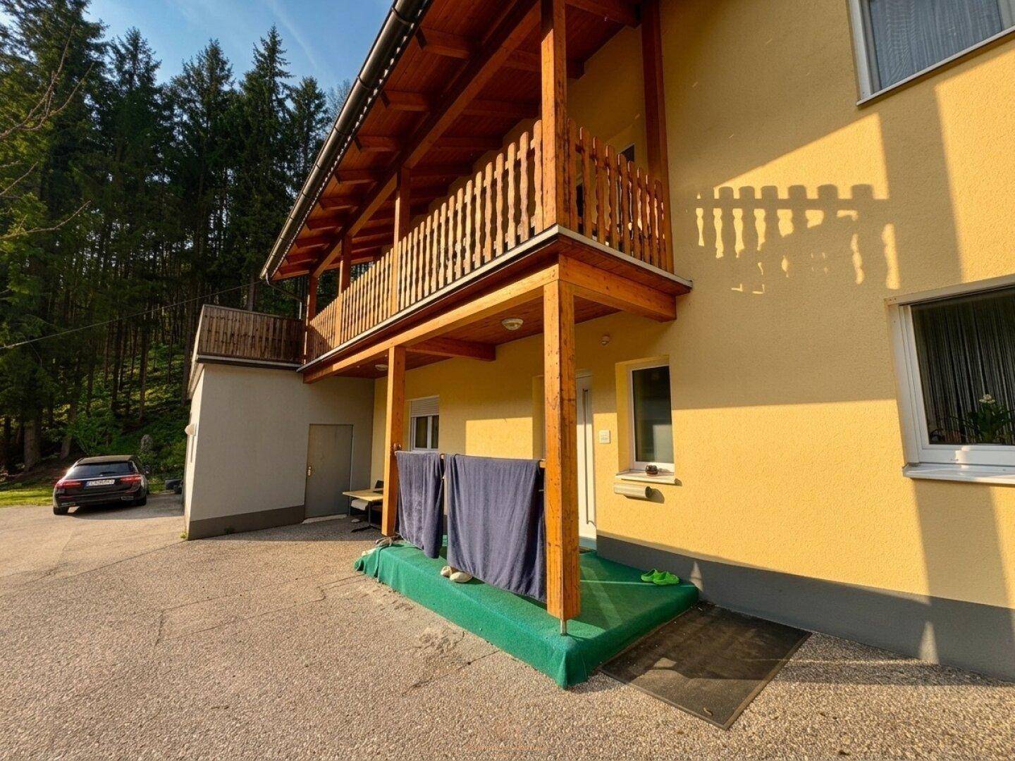 Eingangsbereich des Hauses mit Holzbalkon und Wäscheleinen, umgeben von grüner Natur.